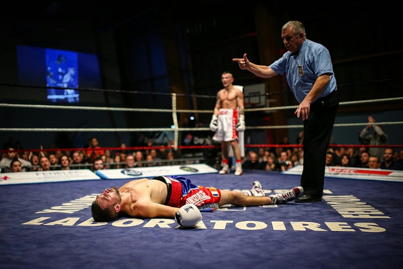 Boxer lying on the canvas after a knockdown as a referee signals in the ring, with the opposing fighter standing in the background.