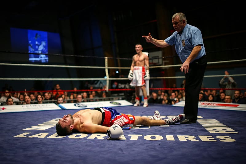 Boxer lying on the canvas after being knocked down during a match as the referee begins the count, with the opponent standing in the background.