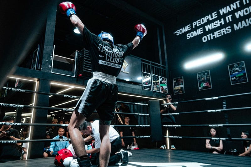 Referee kneels by a knocked down boxer on the canvas while the winning fighter raises his arms in celebration after a stoppage.